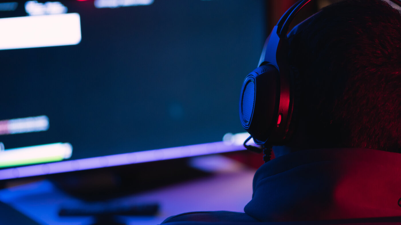 A player seen from behind in a dark room, wearing an over-ear headset lit red and blue, facing a blurred monitor.
